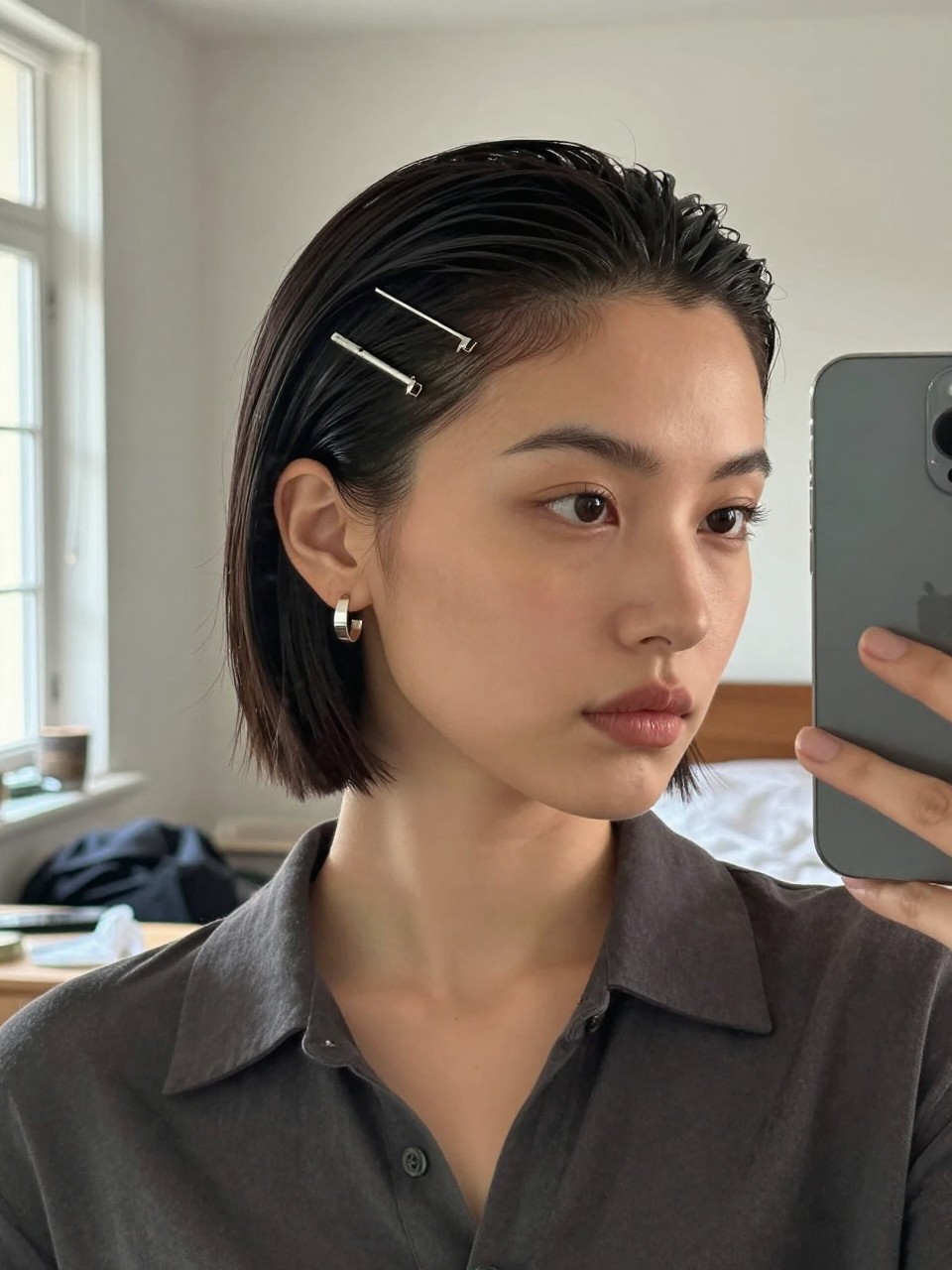 Photo of a woman wearing a sleek slick back short bob with 3 mini hair pins placed on one side near the temple, candid mirror selfie, setting in a sun-drenched bedroom, lighting natural window light, containing a small lived-in detail such as a hair clip clipped to the collar and statement earrings peeking through, latest iPhone photo quality.