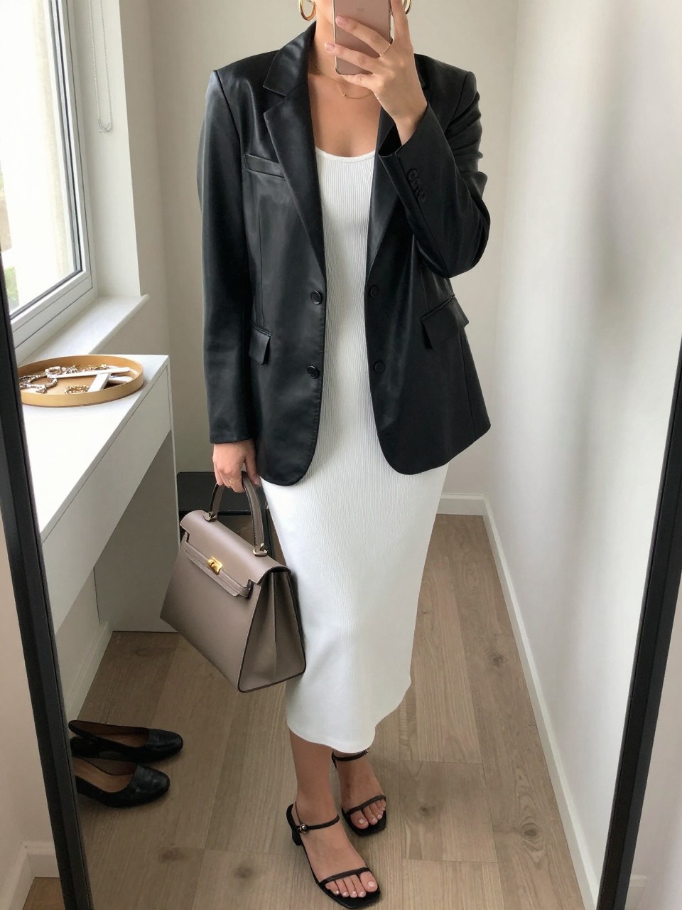 mirror selfie of a woman wearing the exact outfit described above, clearly showing a soft black leather blazer, a white ribbed tank midi dress, black minimal strappy sandals, a taupe top-handle leather bag, and chunky gold hoop earrings, holding a phone in front of a mirror, cropped at NECK so the face does not show, with a relaxed casual selfie pose, in a walk-in closet mirror, natural window light, including jewelry tray on the vanity and pair of heels on the floor nearby, latest iPhone photo quality. (no watermarks on images)