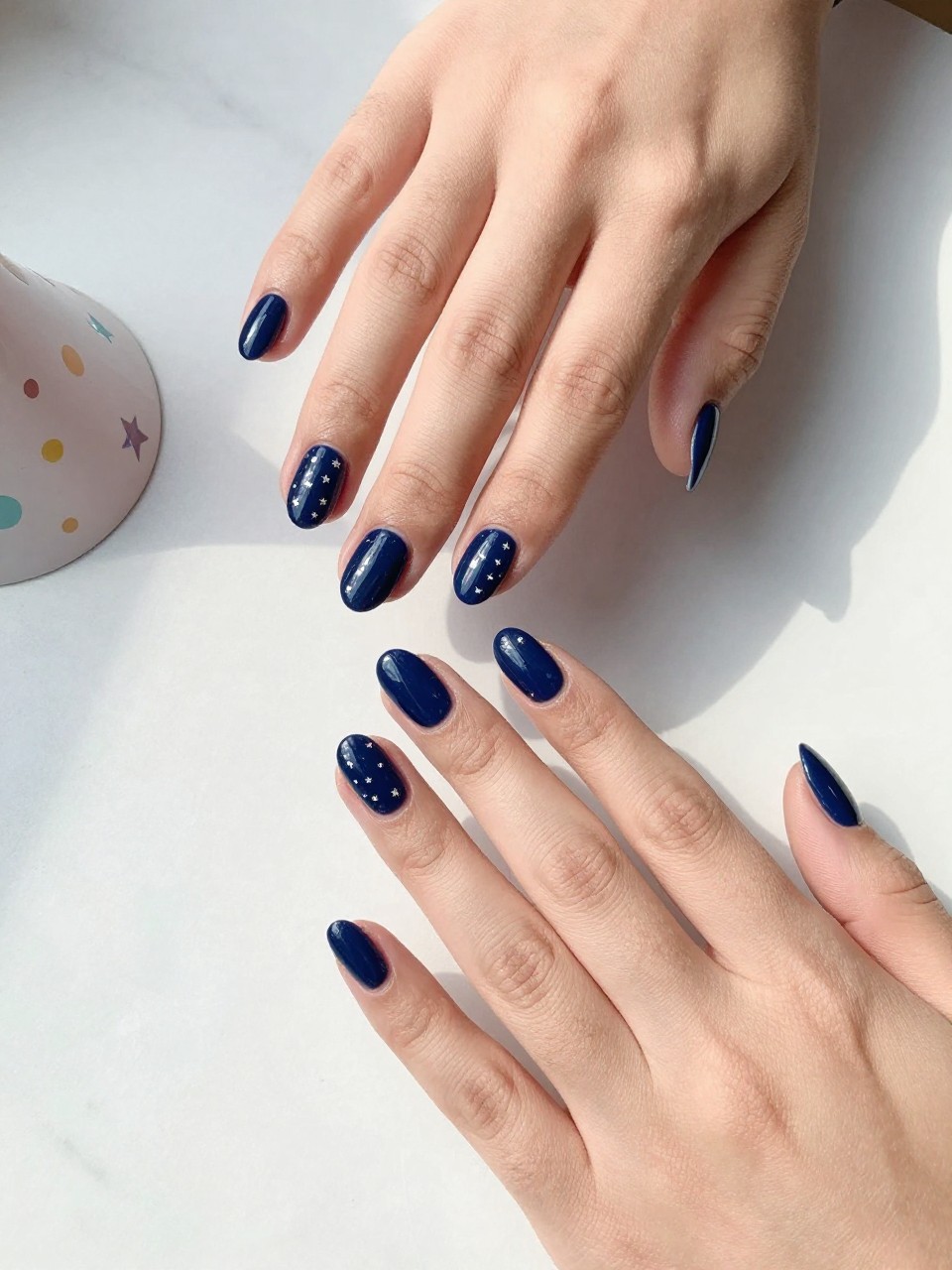 90-degree overhead top-down view of two feminine hands with almond medium nails.
The manicure features a glossy navy blue base with tiny silver star accents scattered across two accent nails.
Include glossy gel finish.

Hands are posed fingers gently spread.

Scene set on a white marble surface with a small party hat nearby.

Bright natural daylight with soft shadows, hyper-realistic lifestyle photography, sharp focus on nails, natural skin texture, realistic hands, no extra fingers.