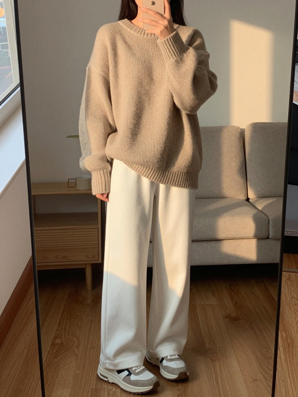 iPhone mirror selfie of a woman in an oversized oatmeal sweater with white wide leg pants and chunky sneakers, relaxed stance holding phone, cropped at NECK so face doesn't show, in a cozy city apartment, evening golden hour, iPhone photo quality.