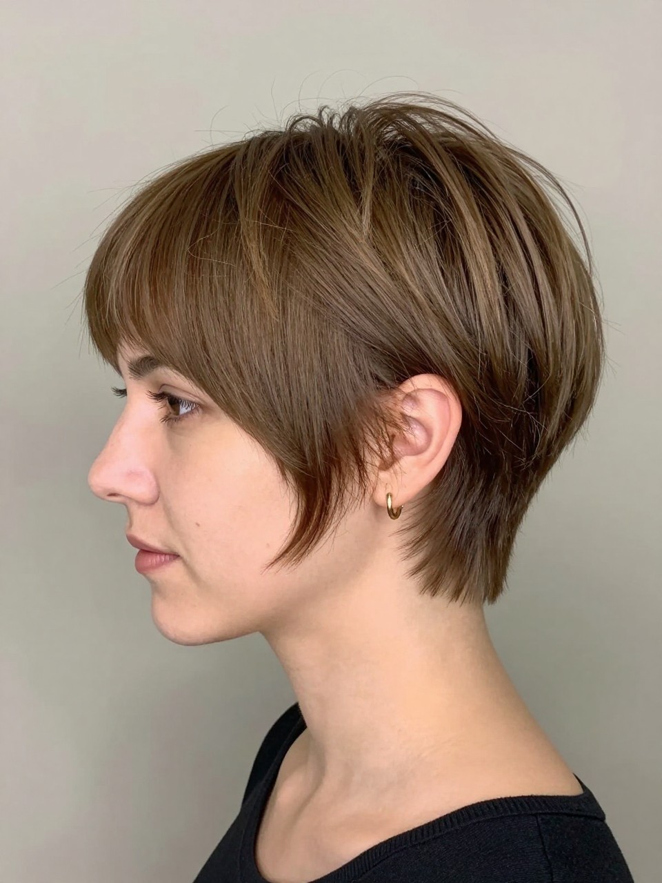 Photo of a white woman wearing an ultra short rounded crop pixie with softly shaped top volume and neat close sides, with the haircut fully visible and clearly defined, shot from a three-quarter angle, in a soft neutral studio, with soft indoor lighting, showing the haircut shape, layers, ends, bangs, and texture clearly, including simple gold earrings, latest iPhone photo quality.