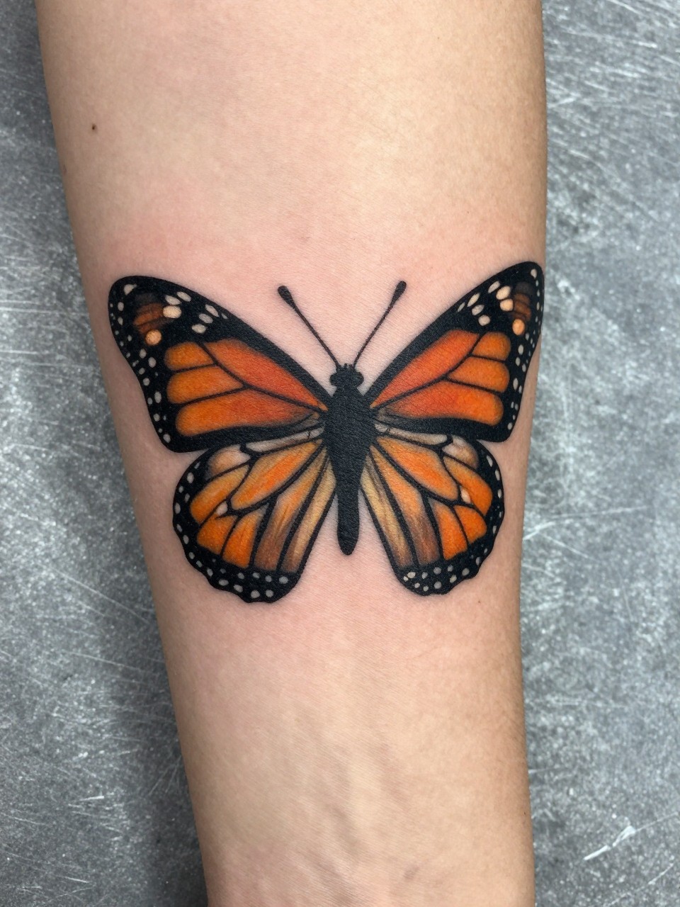 "Close-up of a forearm tattoo: a monarch butterfly with its wings fragmented into clean geometric panels of black and orange. Crisp linework on toned skin, against a brushed steel background."