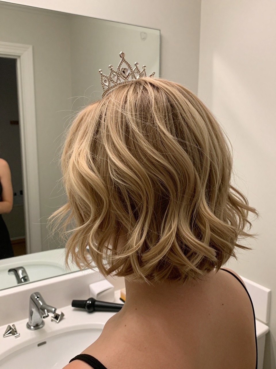 Photo of a white woman wearing short hair with soft waves and a pinned back crown, back-of-head view, setting in a bathroom vanity mirror, lighting soft indoor lighting, containing a small lived-in detail such as bobby pins on the counter and a curling iron on the counter, latest iPhone photo quality.