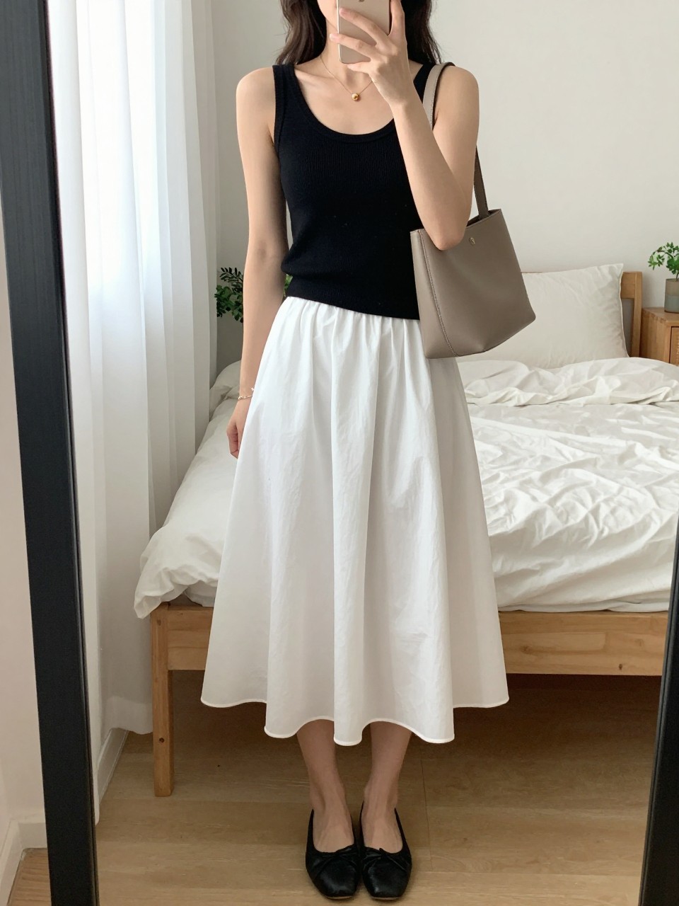 mirror selfie of a woman wearing the exact outfit described above, clearly showing a black fitted knit tank, a white full poplin midi skirt, black two-tone ballet flats, a soft taupe leather shoulder bag, and loose soft waves hairstyle, holding a phone in front of a mirror, cropped at NECK so the face does not show, with a relaxed casual selfie pose, in a feminine-themed bedroom, natural window light, including soft pillows on an unmade bed and small plant on a dresser, latest iPhone photo quality. (no watermarks on images)