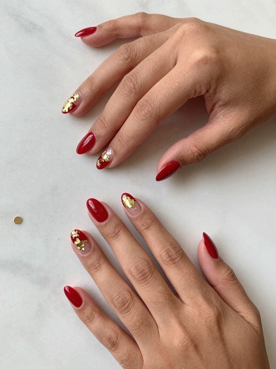 
90-degree overhead top-down view of two feminine black hands with almond medium nails.
The manicure features a glossy crimson red base with scattered gold foil accents on two accent nails.
Include reflective metallic detail with a glossy gel finish.

Hands are posed fingers gently spread.

Scene set on a marble surface with a small gold bracelet nearby.

Bright natural daylight with soft shadows, hyper-realistic lifestyle photography, sharp focus on nails, natural skin texture, realistic hands, no extra fingers.