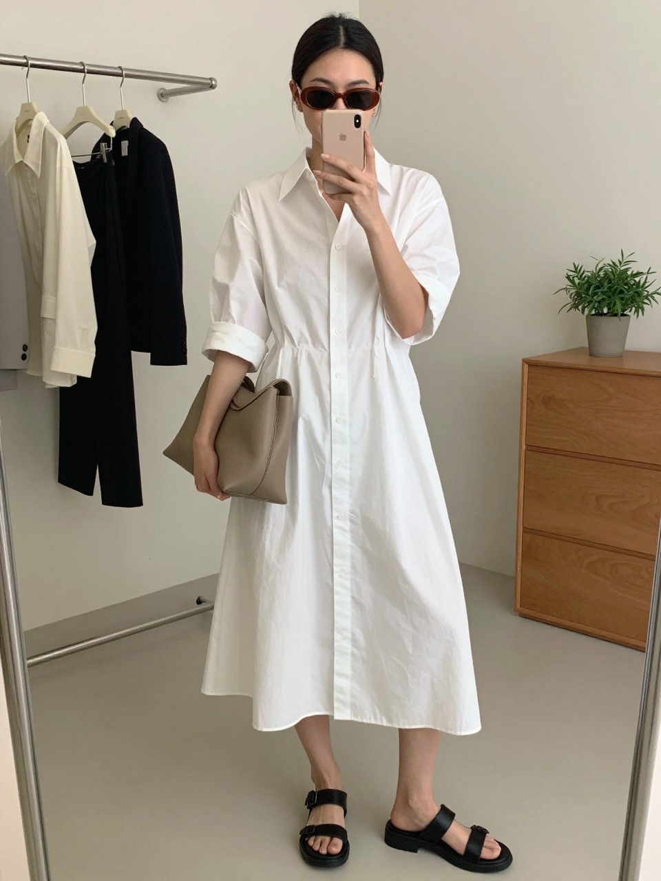 mirror selfie of a woman wearing the exact outfit described above, clearly showing a white cotton poplin midi shirt dress, black chunky leather sandals, a taupe slouchy leather bag, brown oval-frame sunglasses, and hair in a sleek low bun, holding a phone in front of a mirror, cropped at NECK so the face does not show, with a relaxed casual selfie pose, in a fashion studio mirror corner, soft indoor lighting, including clothing rack with garments behind and small plant on a dresser, latest iPhone photo quality. (no watermarks on images)