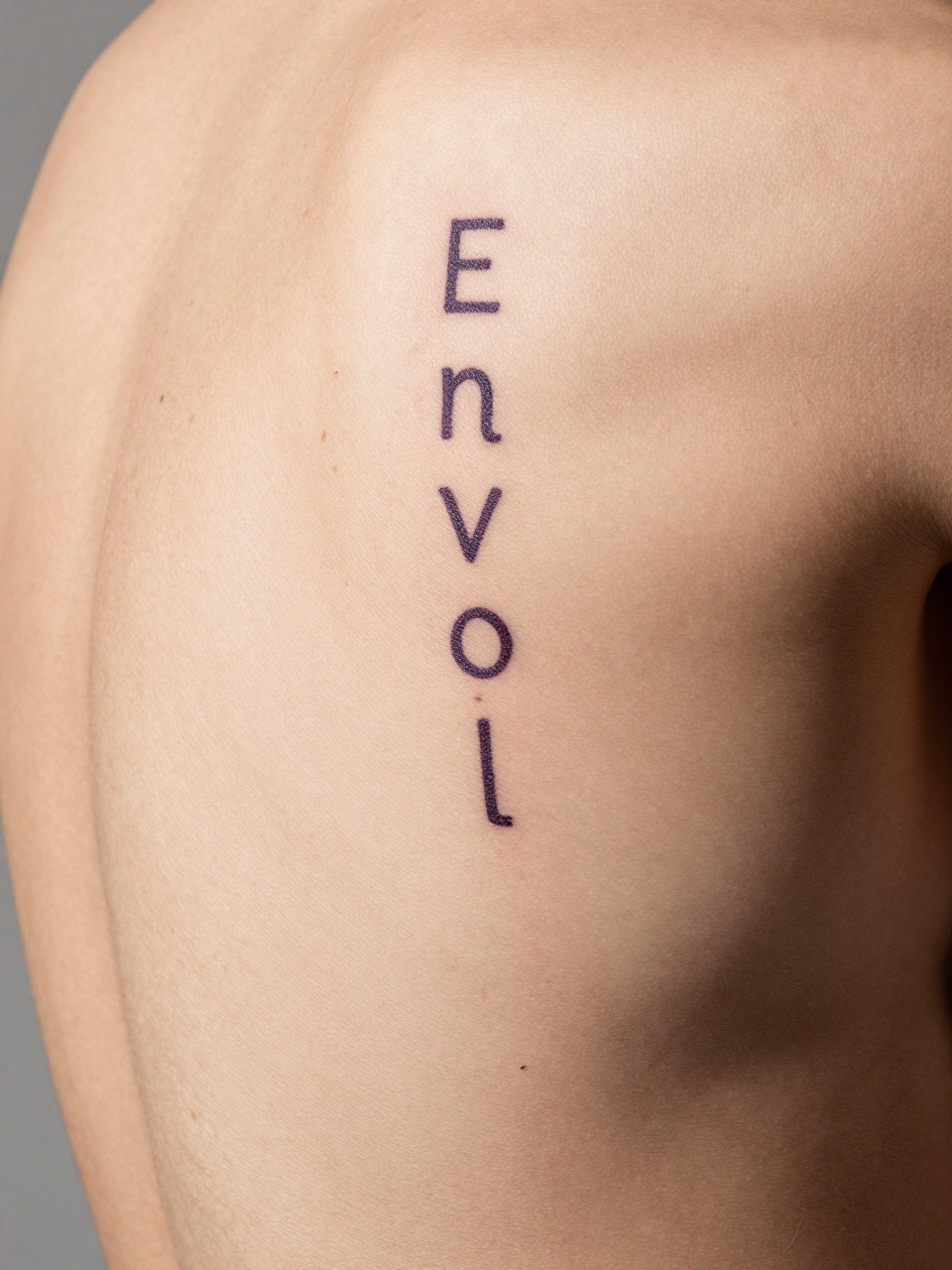 "Detailed shot of 'Envol' in script that looks like it’s trailing upwards, on the side of the ribcage in the skin. Airy and uplifting."