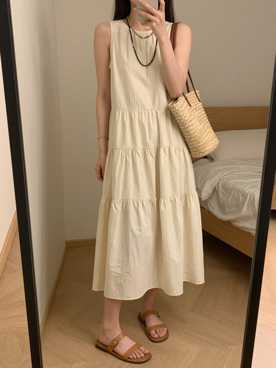 iPhone mirror selfie of a woman wearing a cream tiered layered dress with flat tan leather sandals and a straw market bag, layered beaded necklaces visible, posed with her weight shifted, cropped at NECK so face doesn't show, in a feminine-themed bedroom, warm moody lighting, iPhone photo quality.