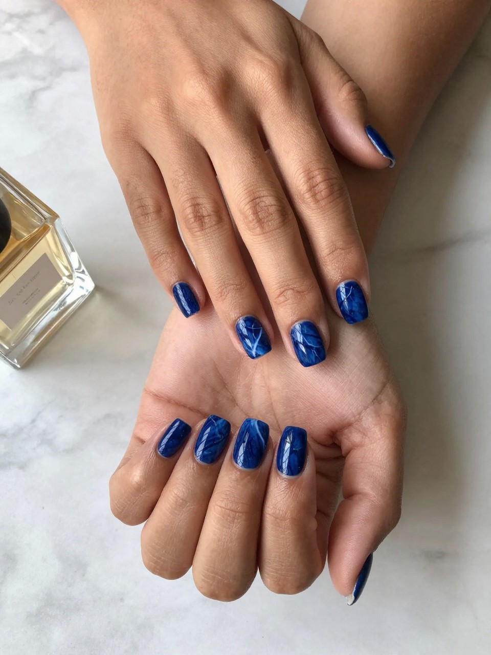 90-degree overhead top-down view of two feminine black hands with rounded square medium nails.
The manicure features a deep blue marble base with soft white vein details running through two accent nails.
Include glossy gel finish.

Hands are posed one hand placed gently over the other.

Scene set on a marble surface with a small perfume bottle nearby.

Bright natural daylight with soft shadows, hyper-realistic lifestyle photography, sharp focus on nails, natural skin texture, realistic hands, no extra fingers.