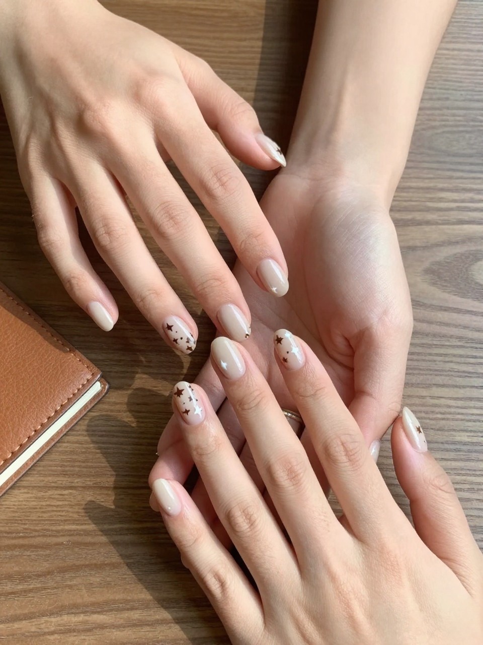 90-degree overhead top-down view of two feminine hands with oval medium nails.
The manicure features a soft beige base with brown and white star accents across the nails.
Include glossy gel finish.

Hands are posed fingers gently spread.

Scene set on a wooden table with a small leather notebook nearby.

Bright natural daylight with soft shadows, hyper-realistic lifestyle photography, sharp focus on nails, natural skin texture, realistic hands, no extra fingers.