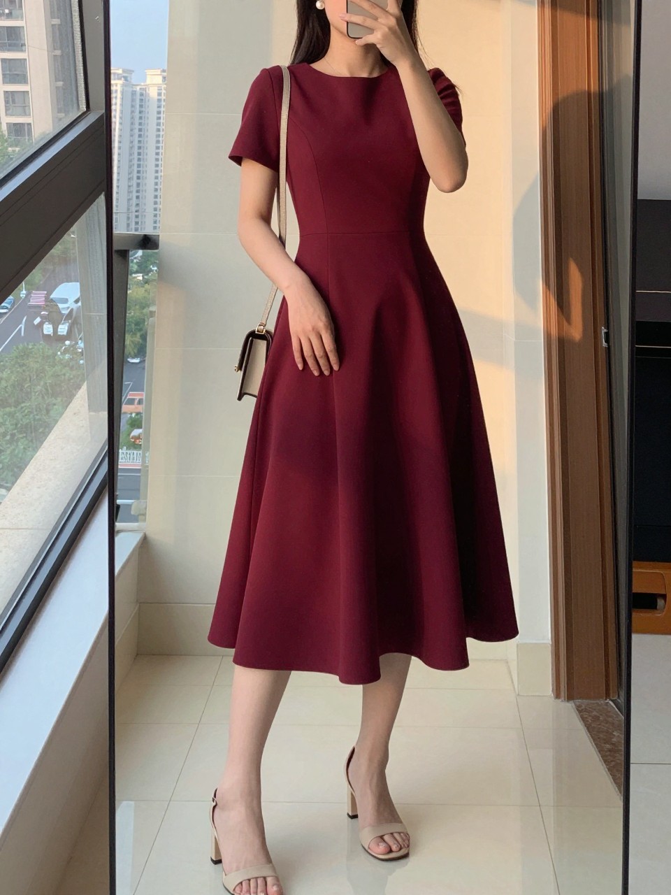 iPhone mirror selfie of a woman wearing a burgundy fit-and-flare dress with a fitted bodice and full knee-length skirt, neutral block heel sandals, and pearl stud earrings, holding a small crossbody bag, posed with one hand gently on the skirt, cropped at NECK so face doesn't show, in a city apartment, evening golden hour, iPhone photo quality.
