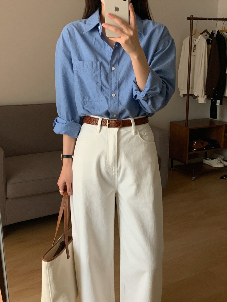 iPhone mirror selfie of a woman in a chambray shirt tucked into white wide leg pants with a woven belt, holding a tote, standing straight with phone down, cropped at NECK so face doesn't show, in a city apartment, warm moody lighting, iPhone photo quality.
