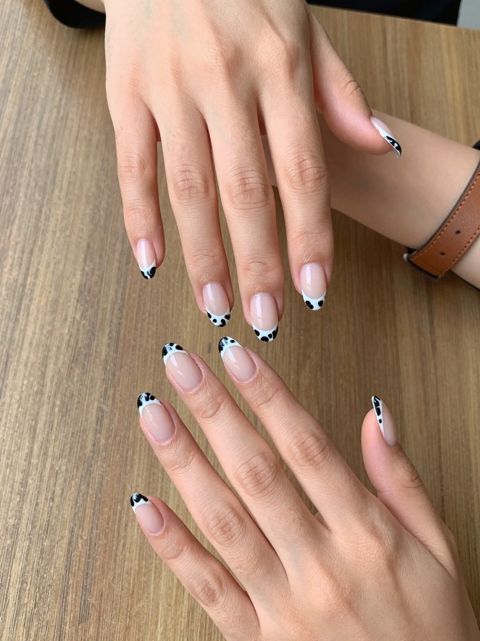 90-degree overhead top-down view of two feminine black hands with almond medium nails.
The manicure features a nude base with black cow print French tips along the edges.
Include glossy gel finish.

Hands are posed fingers gently spread.

Scene set on a wooden table with a leather belt nearby.

Bright natural daylight with soft shadows, hyper-realistic lifestyle photography, sharp focus on nails, natural skin texture, realistic hands, no extra fingers.