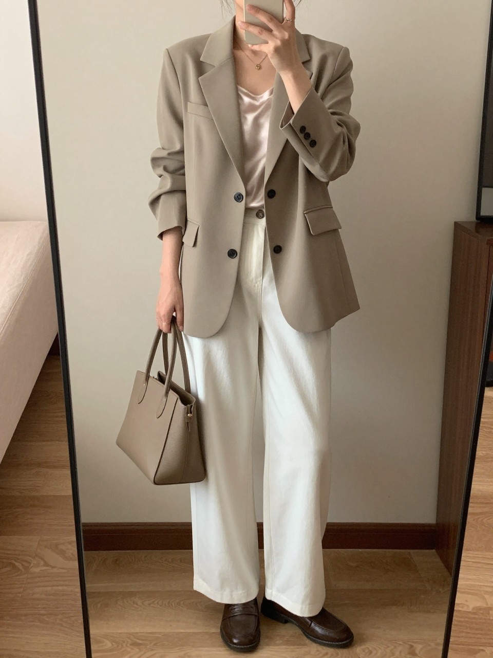 iPhone mirror selfie of a woman in a beige blazer over a silk top with white wide leg pants and loafers, holding a structured bag, standing straight with phone down, cropped at NECK so face doesn't show, in a minimal city apartment, warm moody lighting, iPhone photo quality.