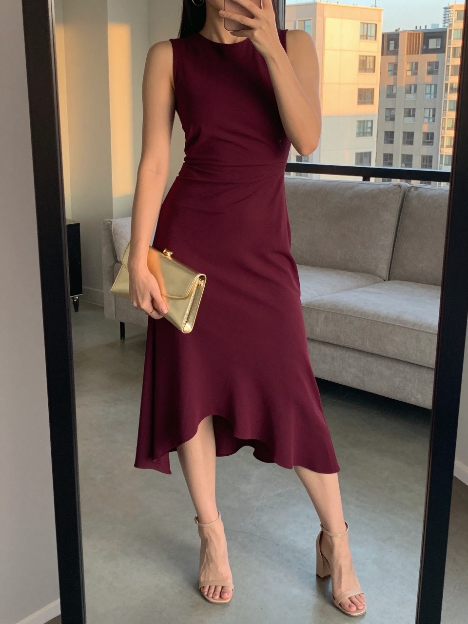 iPhone mirror selfie of a woman wearing a burgundy asymmetrical hem dress with nude ankle strap block heel sandals and a gold metallic clutch, hoop earrings visible, posed to show the hemline, cropped at NECK so face doesn't show, in a city apartment, evening golden hour, iPhone photo quality.