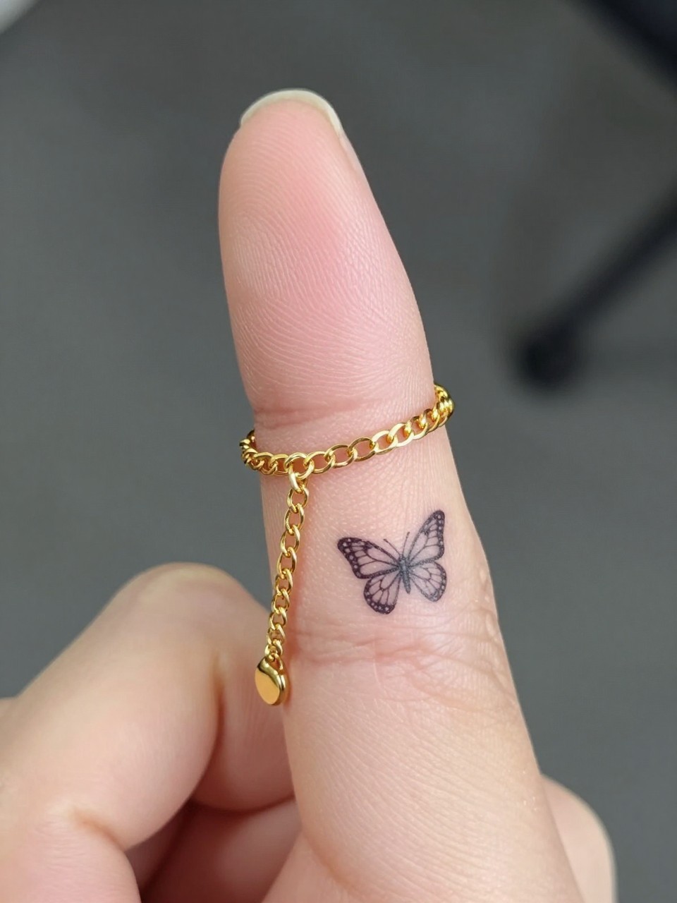 "Extreme close-up of a pinky finger with a fine chain tattoo encircling it, a micro butterfly tattoo dangling off the side. The hand is touching a delicate gold real chain for texture contrast."