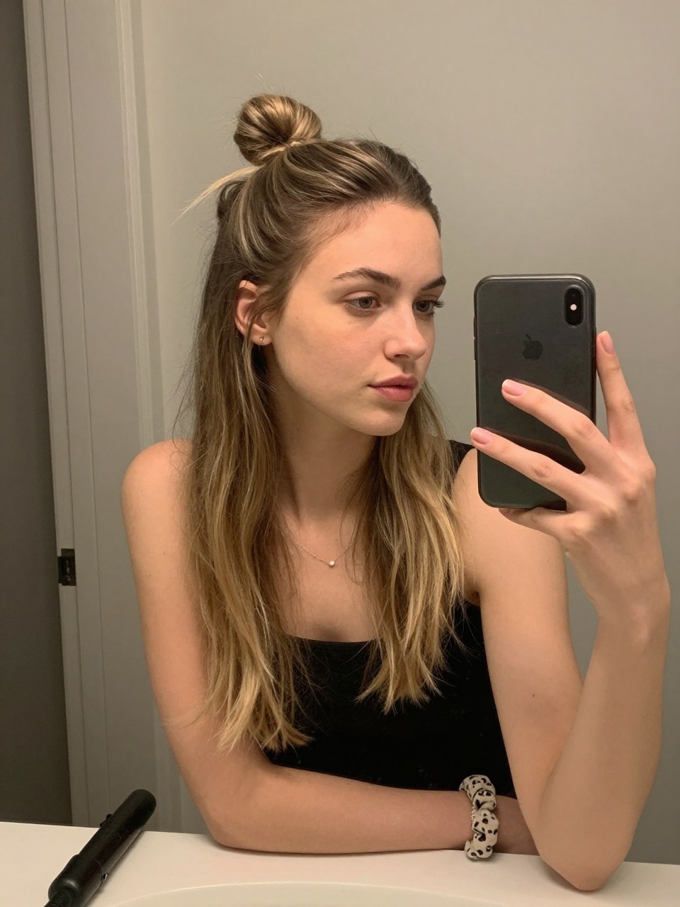 Photo of a white woman wearing a half-up mini bun with the rest of the hair down, candid mirror selfie, setting in a bathroom vanity mirror, lighting soft indoor lighting, containing a small lived-in detail such as a curling iron on the counter and a scrunchie on the wrist, latest iPhone photo quality.