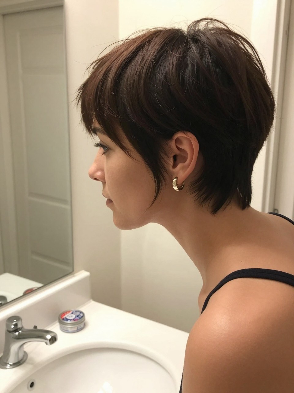 Photo of a woman wearing a polished pixie with a soft side sweep, side profile view, setting in a bathroom vanity mirror, lighting soft indoor lighting, containing a small lived-in detail such as a small pomade tin on the counter and statement earrings peeking through, latest iPhone photo quality.