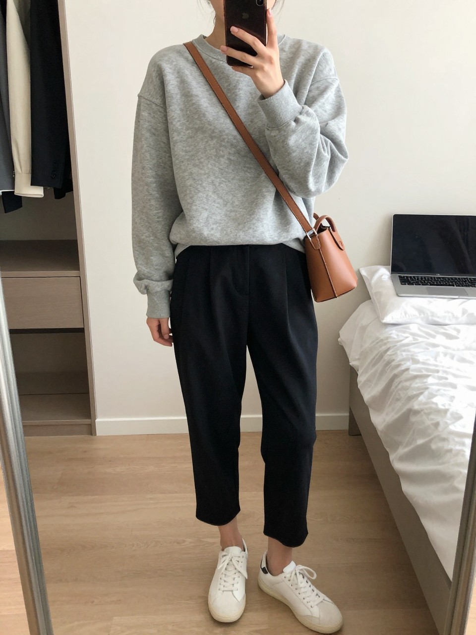 mirror selfie of a woman wearing the exact outfit described above, clearly showing a heather gray cotton sweatshirt, black pleated ankle-length trousers, white leather sneakers, a camel crossbody leather bag, and slim tortoise shell sunglasses, holding a phone in front of a mirror, cropped at NECK so the face does not show, with a relaxed casual selfie pose, in a minimal dressing room, soft indoor lighting, including laptop or tablet on the bed and open closet with clothes visible, latest iPhone photo quality. (no watermarks on images)