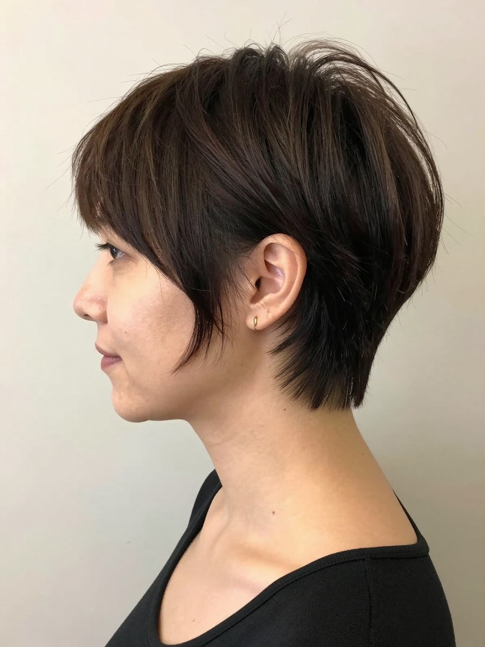 Photo of a white woman wearing a tapered pixie with close sides, a neat neckline, and lifted top layers, with the haircut fully visible and clearly defined, shot from a side profile, in a soft neutral studio, with soft indoor lighting, showing the haircut shape, layers, ends, bangs, and texture clearly, including simple gold earrings, latest iPhone photo quality.