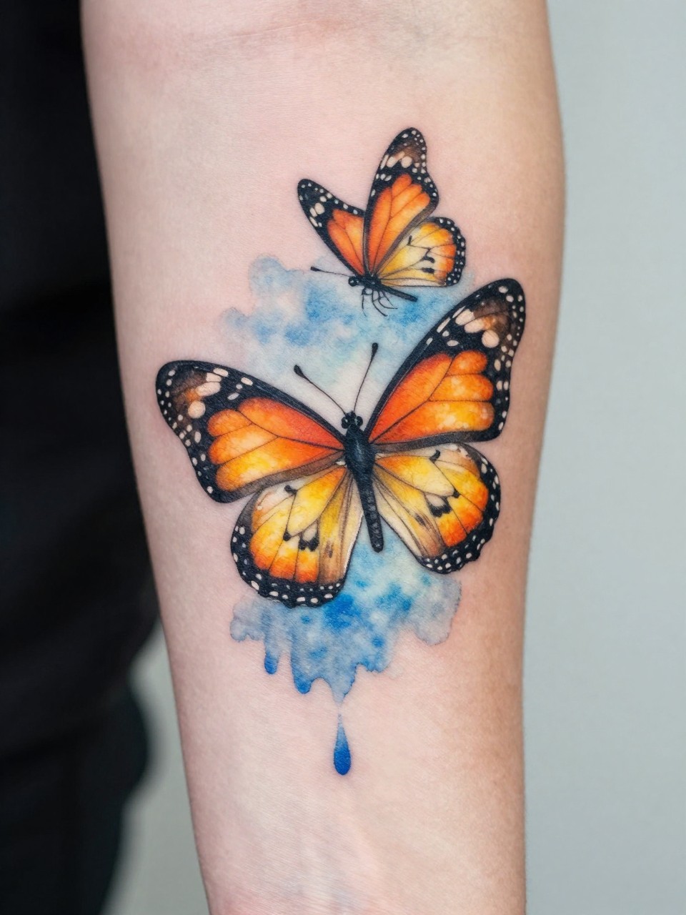 "Close-up macro shot of a watercolor tattoo on a forearm: a monarch butterfly with soft orange and yellow bleeds, blending into a blue wash that transitions into a second butterfly above. The skin is clean and the colors are luminous."