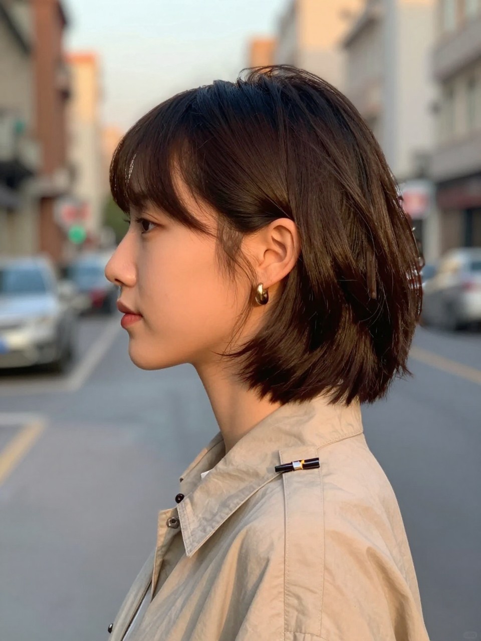 Photo of a woman wearing a soft blowout lob with both sides tucked behind the ears, side profile view, setting in a softly blurred city street background, lighting golden hour glow, containing a small lived-in detail such as statement earrings peeking through and a hair clip clipped to the collar, latest iPhone photo quality.