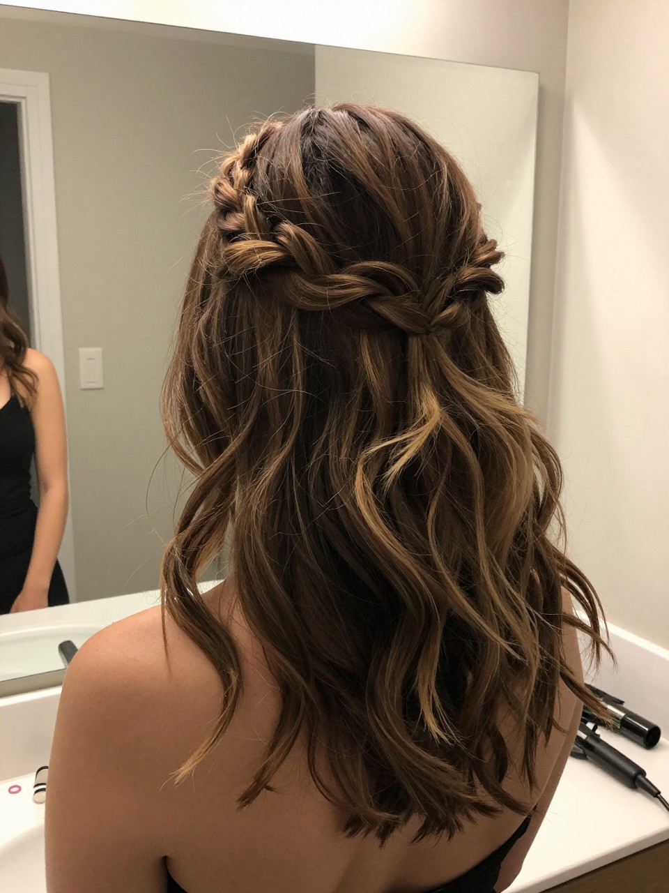 Photo of a woman wearing a half-up braided crown with loose waves on medium-length hair, back-of-head view, setting in a bathroom vanity mirror, lighting soft indoor lighting, containing a small lived-in detail such as mini elastics on the counter and a curling iron nearby, latest iPhone photo quality.