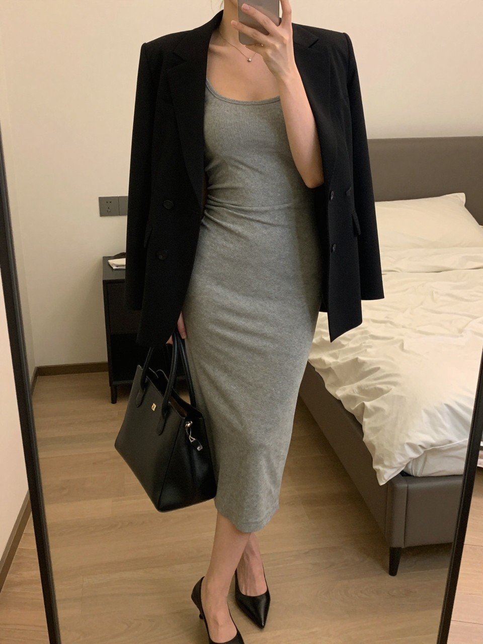 iPhone mirror selfie of a woman wearing a grey bodycon dress with a black structured blazer over it and black pointed-toe heels, holding a black leather tote, posed with one hand in her blazer pocket, cropped at NECK so face doesn't show, in a feminine-themed bedroom, warm moody lighting, iPhone photo quality.