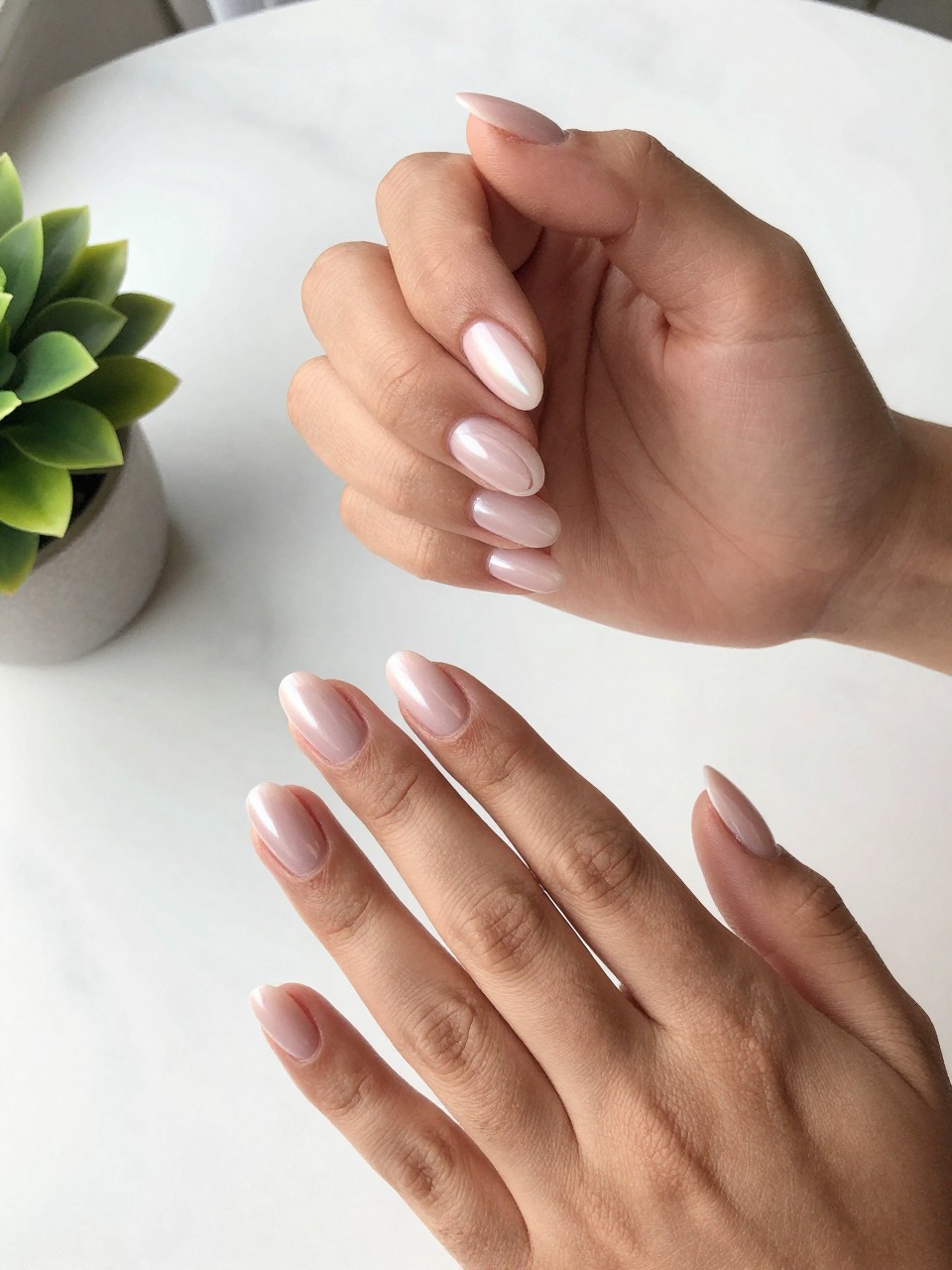 90-degree overhead top-down view of two feminine black hands with almond medium nails.
The manicure features a milky pink chrome base with curved negative space details near the cuticle on two accent nails.
Include glossy reflective shine with a gel finish.

Hands are posed fingers gently spread.

Scene set on a white marble surface with a small plant nearby.

Bright natural daylight with soft shadows, hyper-realistic lifestyle photography, sharp focus on nails, natural skin texture, realistic hands, no extra fingers.