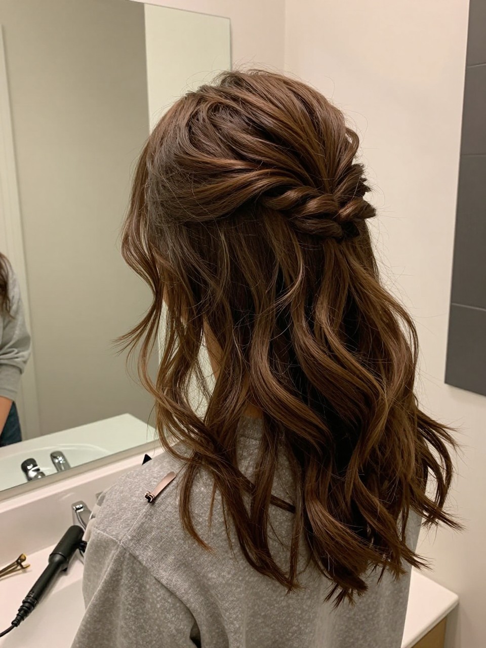 Photo of a woman wearing a half-up twisted crown with loose waves on medium-length hair, back-of-head view, setting in a bathroom vanity mirror, lighting soft indoor lighting, containing a small lived-in detail such as a curling iron on the counter and a hair clip clipped to the collar, latest iPhone photo quality.