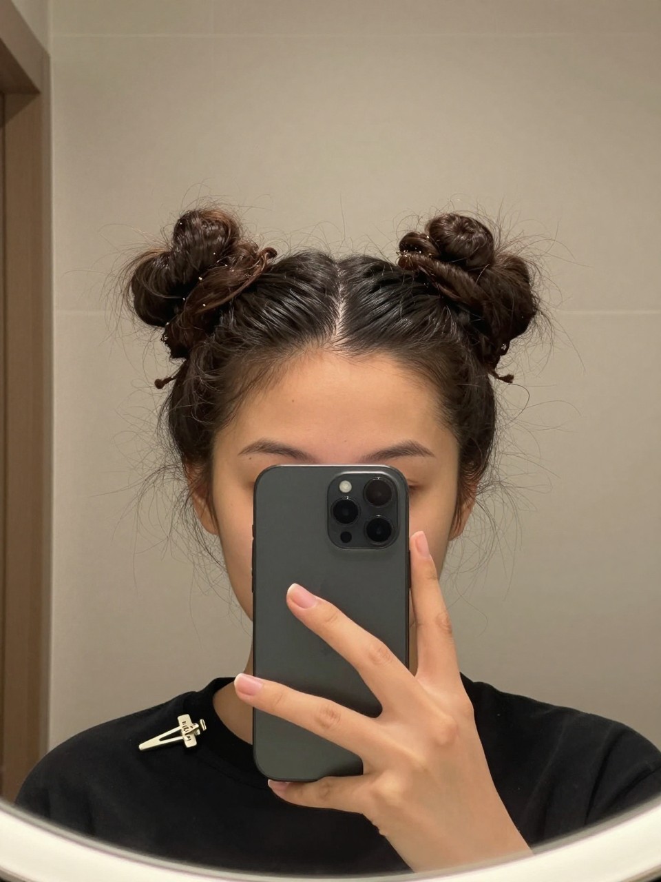 Photo of a woman wearing curly space buns, front-facing mirror selfie showing both buns at the crown, setting is a bathroom vanity mirror with soft indoor lighting, containing a hair clip clipped to her collar, iPhone photo quality.
