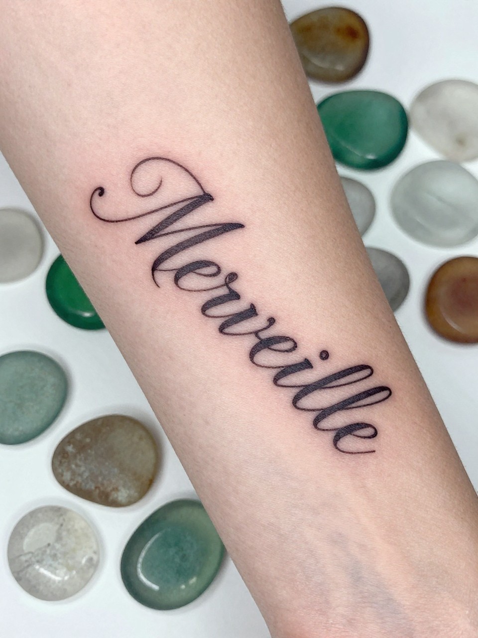"Macro photograph of 'Merveille' in elegant, fine-line script on the inner arm. The arm brushes against a collection of intriguing, smooth sea glass. Wondrous, light-filled detail."
