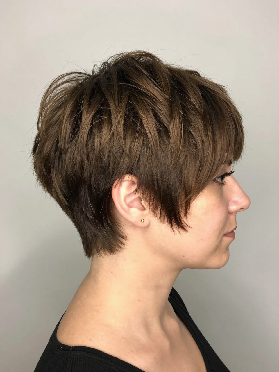 Photo of a white american woman wearing a choppy crown pixie with tapered sides and textured top layers, with the haircut fully visible and clearly defined, shot from a side profile, in a soft neutral studio, with soft indoor lighting, showing the haircut shape, layers, ends, bangs, and texture clearly, including simple gold earrings, latest iPhone photo quality.