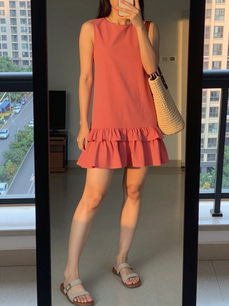 iPhone mirror selfie of a woman wearing a coral dress with a ruffled hem and flat neutral sandals, a woven tote bag over her arm, a thin gold necklace visible, posed with her weight shifted to one leg, cropped at NECK so face doesn't show, in a city apartment, evening golden hour, iPhone photo quality.