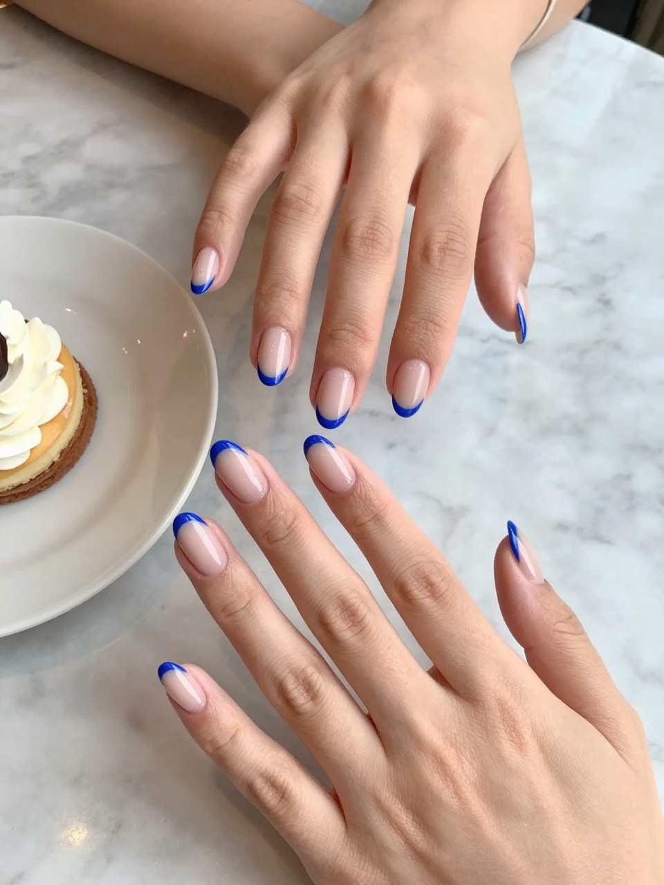 90-degree overhead top-down view of two feminine hands with almond medium nails.
The manicure features a soft nude base with bold cobalt blue French tips along the edges.
Include glossy gel finish.

Hands are posed fingers gently spread.

Scene set on a marble café table with a small dessert plate nearby.

Bright natural daylight with soft shadows, hyper-realistic lifestyle photography, sharp focus on nails, natural skin texture, realistic hands, no extra fingers.
