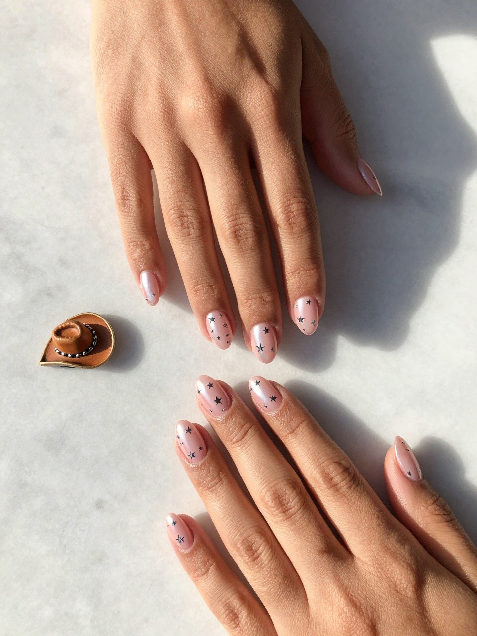 90-degree overhead top-down view of two feminine black hands with almond medium nails.
The manicure features a pink chrome base with tiny silver star accents scattered across two nails.
Include reflective glossy gel finish.

Hands are posed fingers gently spread.

Scene set on a marble surface with a small cowboy hat charm nearby.

Bright natural daylight with soft shadows, hyper-realistic lifestyle photography, sharp focus on nails, natural skin texture, realistic hands, no extra fingers.