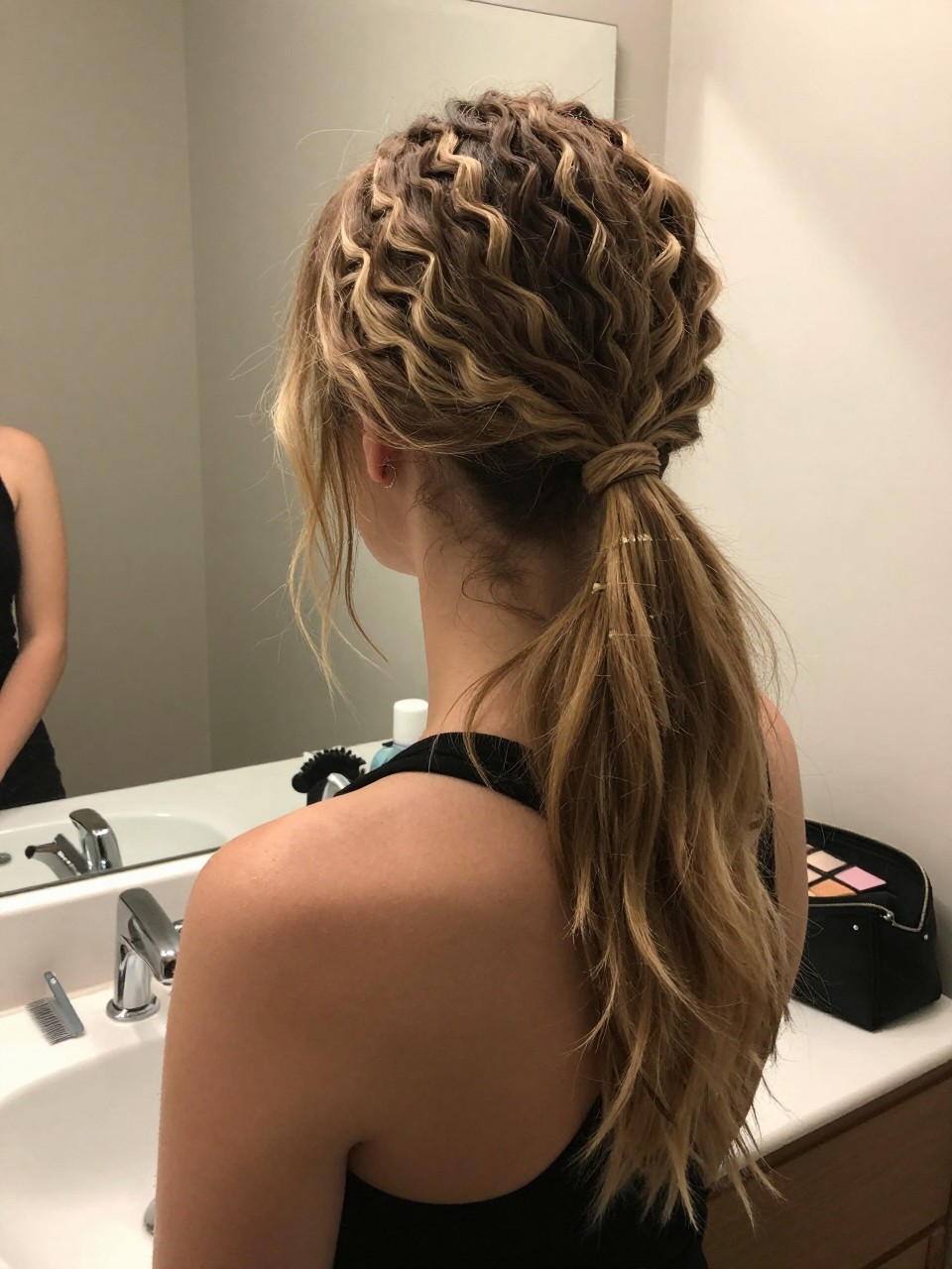 Photo of a white woman wearing a zigzag part with a loose low ponytail, back-of-head view, setting in a bathroom vanity mirror, lighting soft indoor lighting, containing a small lived-in detail such as a rat-tail comb on the counter and a makeup bag nearby, latest iPhone photo quality.