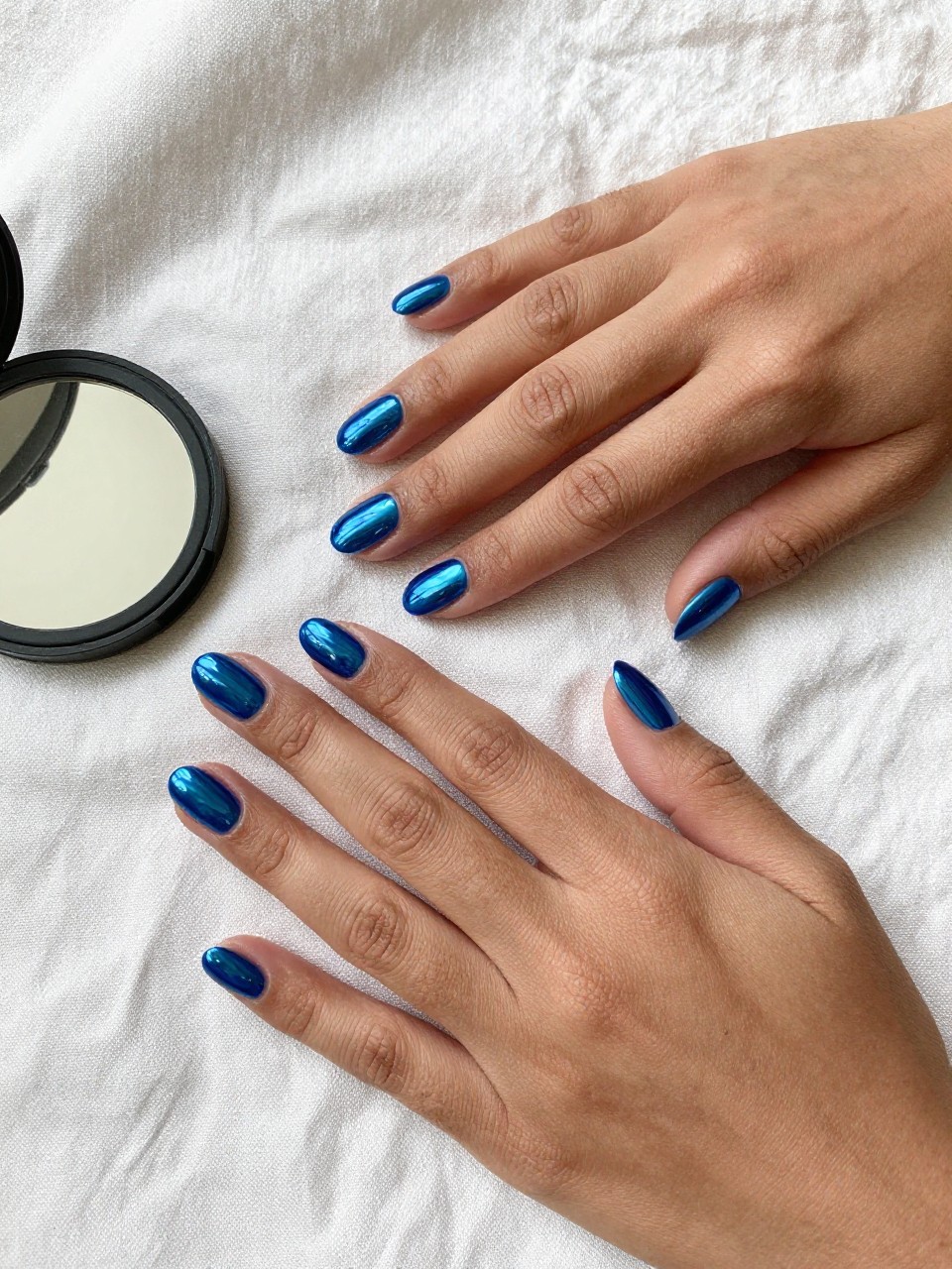 90-degree overhead top-down view of two feminine black hands with oval medium nails.
The manicure features a full blue chrome base with a mirror-like reflective finish across all nails.
Include smooth glossy gel finish.

Hands are posed one hand placed over the other.

Scene set on a soft linen cloth with a small compact mirror nearby.

Bright natural daylight with soft shadows, hyper-realistic lifestyle photography, sharp focus on nails, natural skin texture, realistic hands, no extra fingers.
