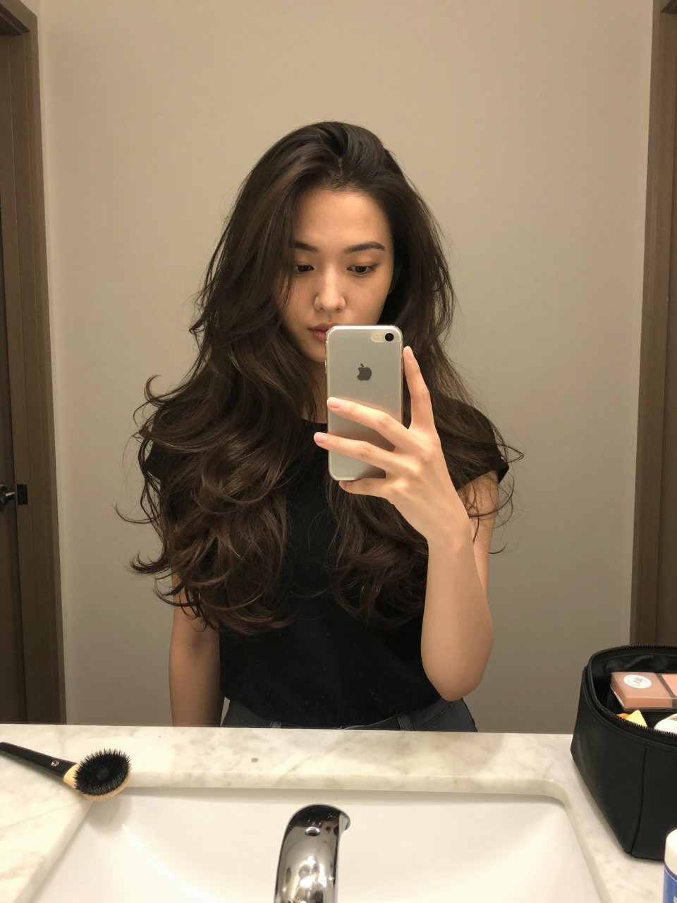 Photo of a woman wearing a classic blowout with bouncy ends on long hair, candid mirror selfie, setting in a bathroom vanity mirror, lighting soft indoor lighting, containing a small lived-in detail such as a round brush on the counter and a makeup bag nearby, latest iPhone photo quality.