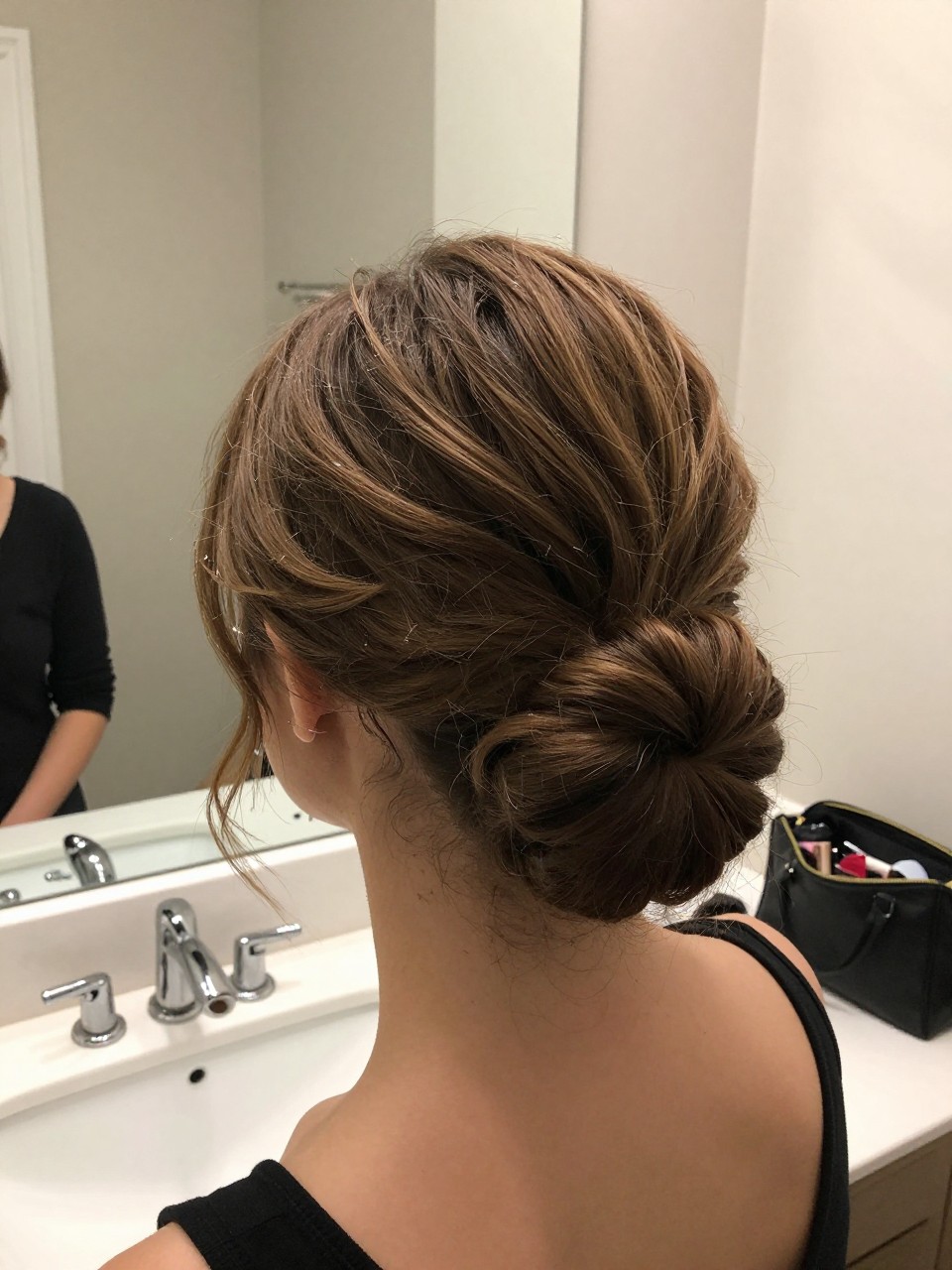Photo of a woman wearing a textured low chignon on a short lob, back-of-head view to show pinned bun shape, setting in a bathroom vanity mirror, lighting soft indoor lighting, containing a small lived-in detail such as bobby pins on the counter and a makeup bag nearby, latest iPhone photo quality.