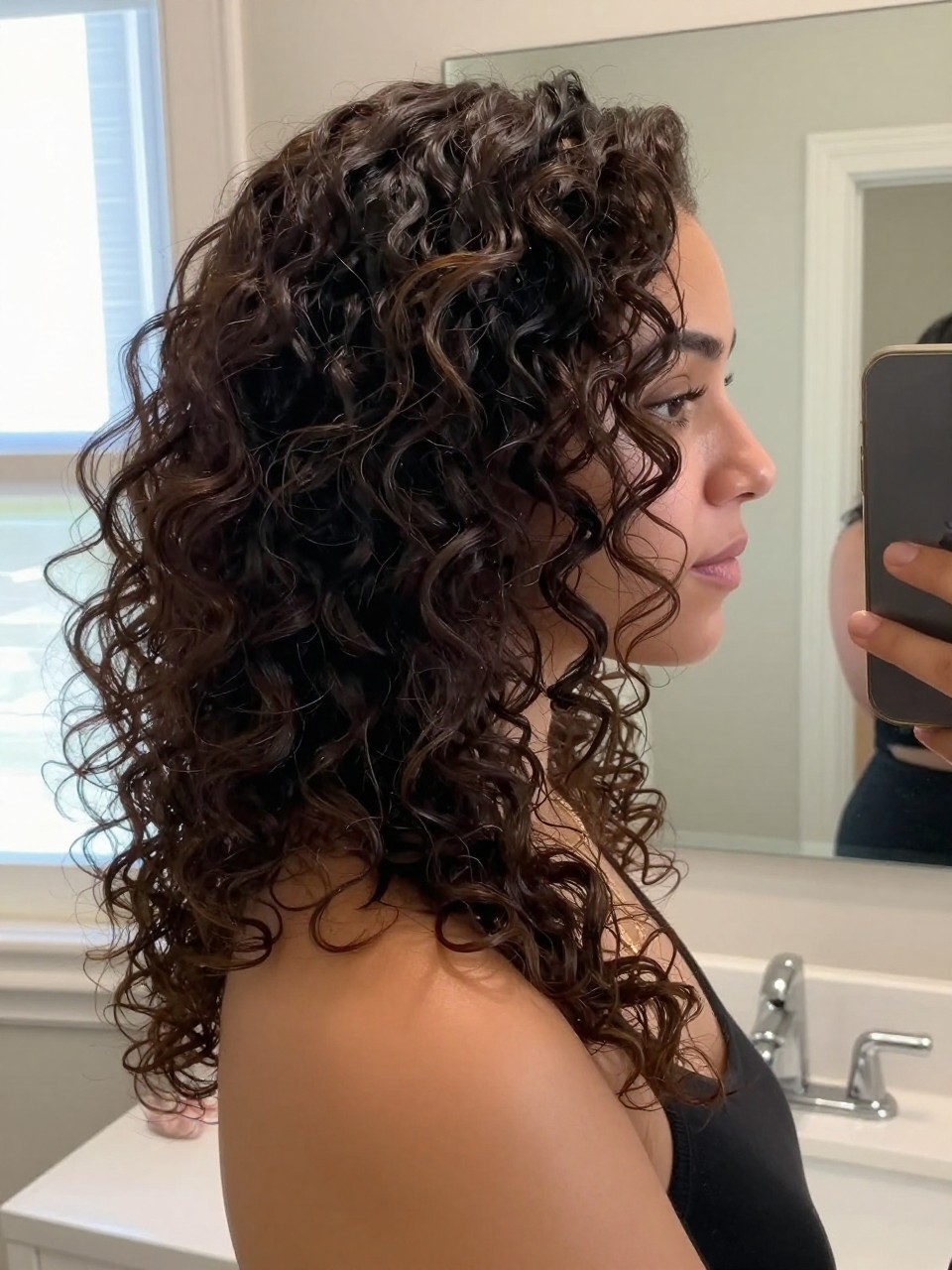 Photo of a woman wearing a deep side part with cascading curls, side profile view showing curls draped over one shoulder, setting is a bathroom vanity mirror with natural window light, containing a simple gold necklace visible, iPhone photo quality.