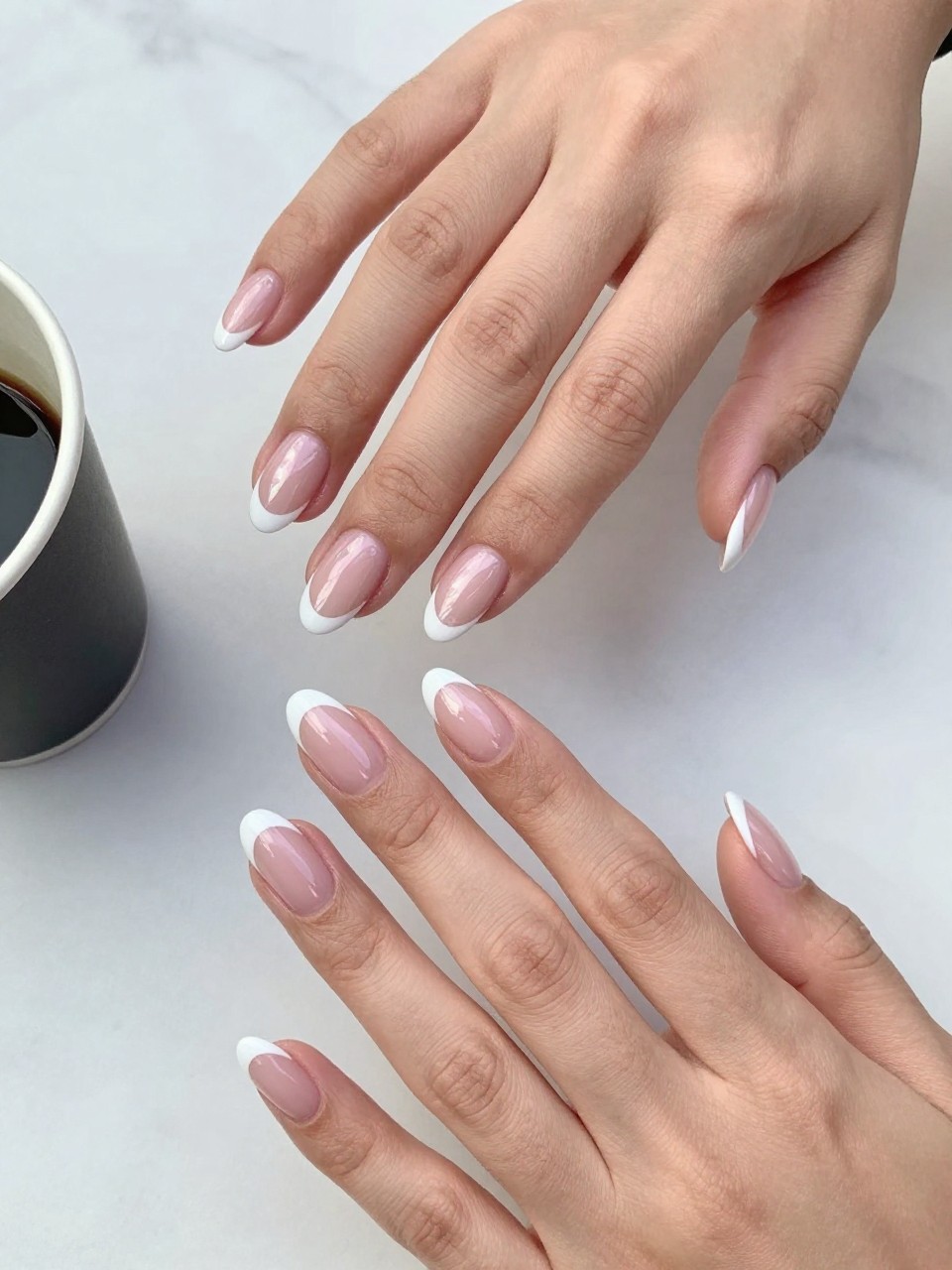 90-degree overhead top-down view of two feminine hands with almond medium nails.
The manicure features a milky pink chrome base with ultra-thin white micro French tips along the edges.
Include delicate chrome reflection across the nails with a glossy gel finish.

Hands are posed with fingers gently spread.

Scene set on a white marble surface with a small coffee cup nearby.

Bright natural daylight with soft shadows, hyper-realistic lifestyle photography, sharp focus on nails, natural skin texture, realistic hands, no extra fingers.