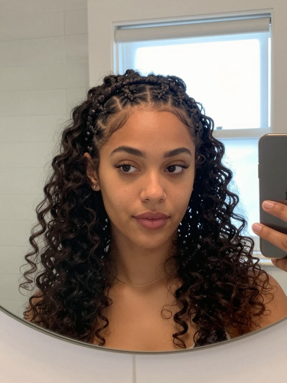 Photo of a woman wearing a curly braided headband, front-facing view showing the braid across the top of her head, setting is a bathroom vanity mirror with natural window light, containing a simple gold necklace visible, iPhone photo quality.