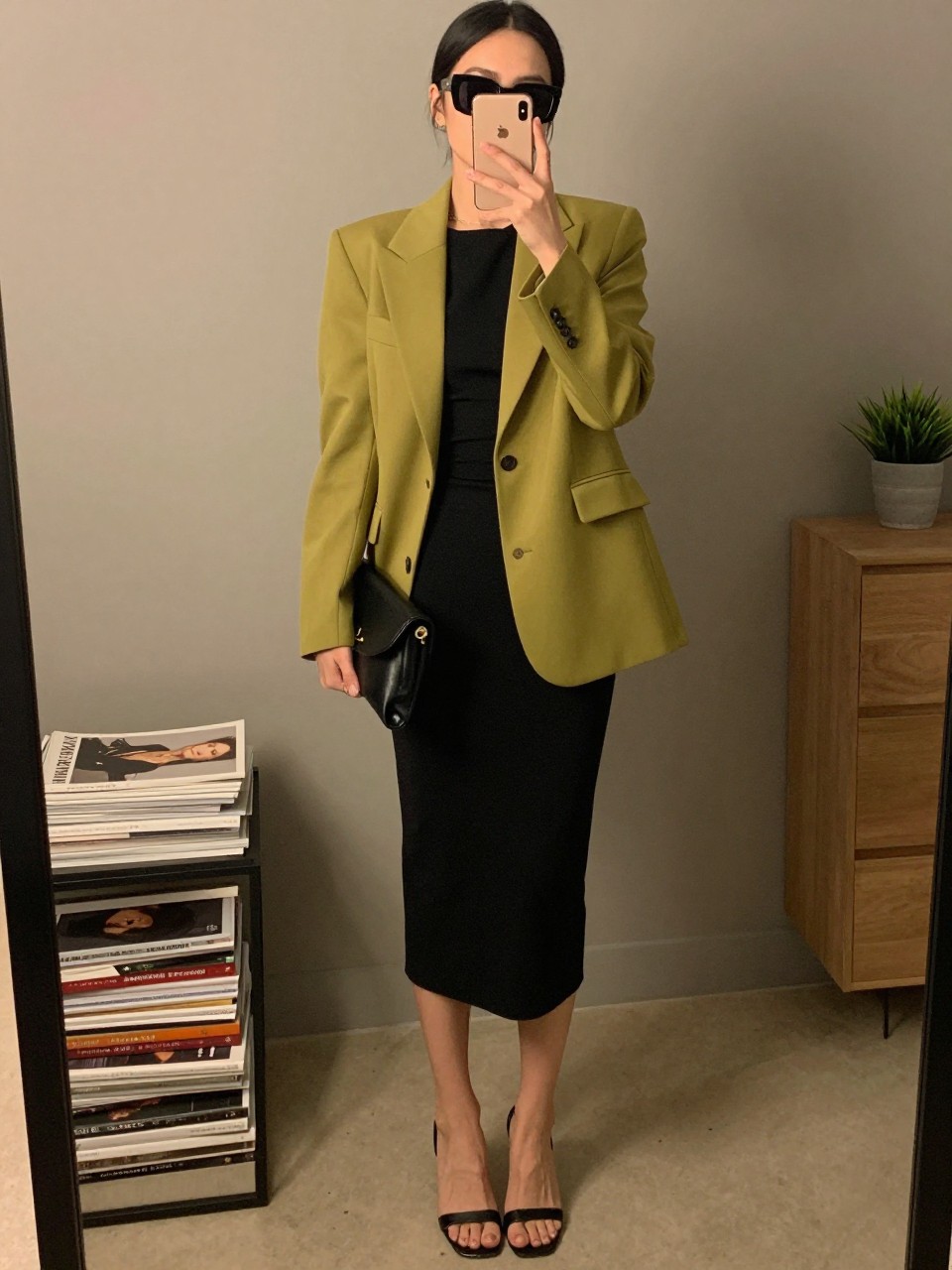 mirror selfie of a woman wearing the exact outfit described above, clearly showing a chartreuse tailored blazer, a black fitted column midi dress, black open-toe heels, a black leather clutch, and oversized black-frame sunglasses, holding a phone in front of a mirror, cropped at NECK so the face does not show, with a relaxed casual selfie pose, in a fashion studio mirror corner, warm moody lighting, including stack of fashion magazines on a side table and small plant on a dresser, latest iPhone photo quality. (no watermarks on images)