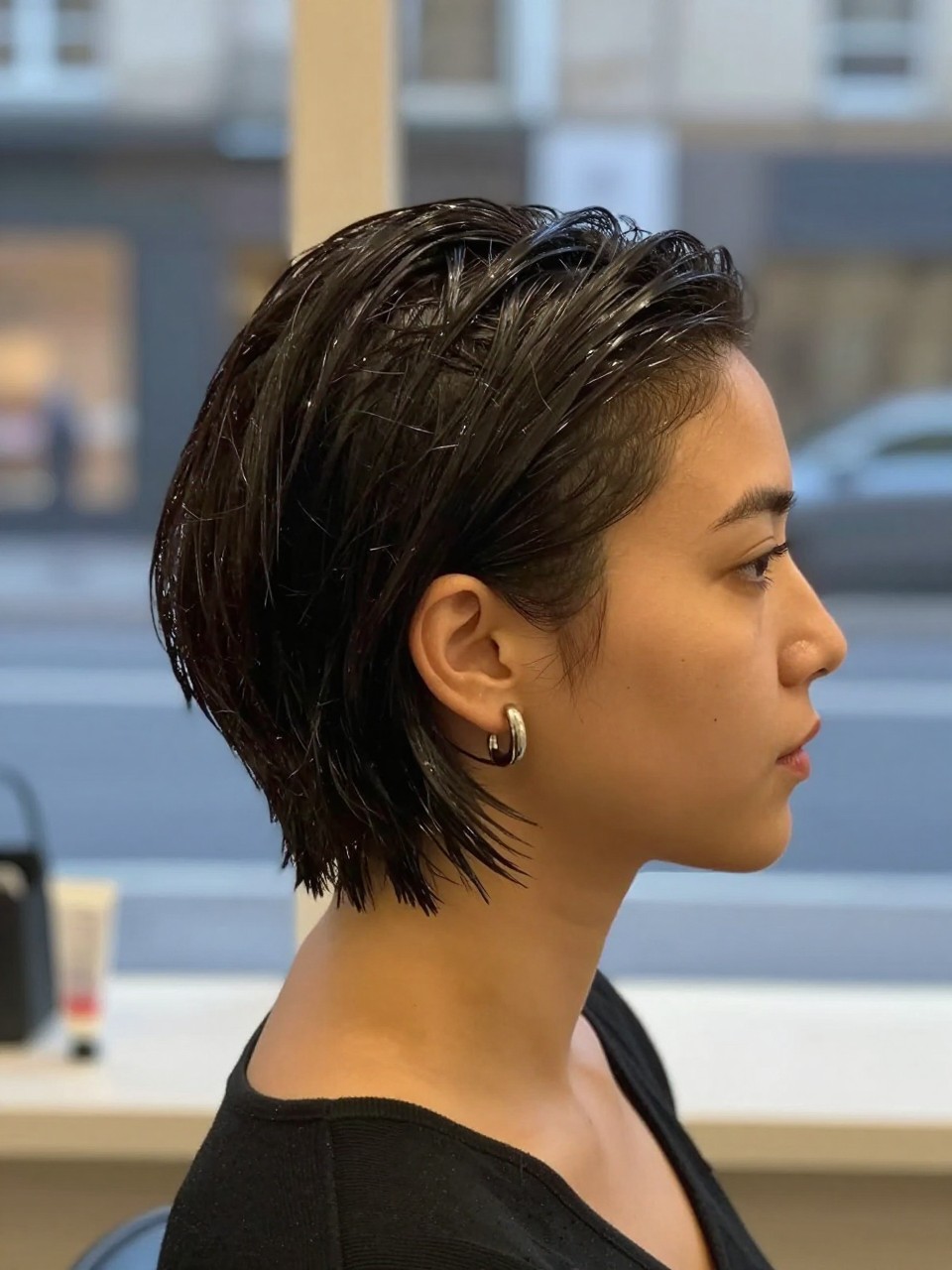 Photo of a woman wearing a wet-look tucked short bob combed back, side profile view, setting in a softly blurred city street background, lighting soft indoor lighting, containing a small lived-in detail such as a small gel tube on the counter and statement earrings peeking through, latest iPhone photo quality.
