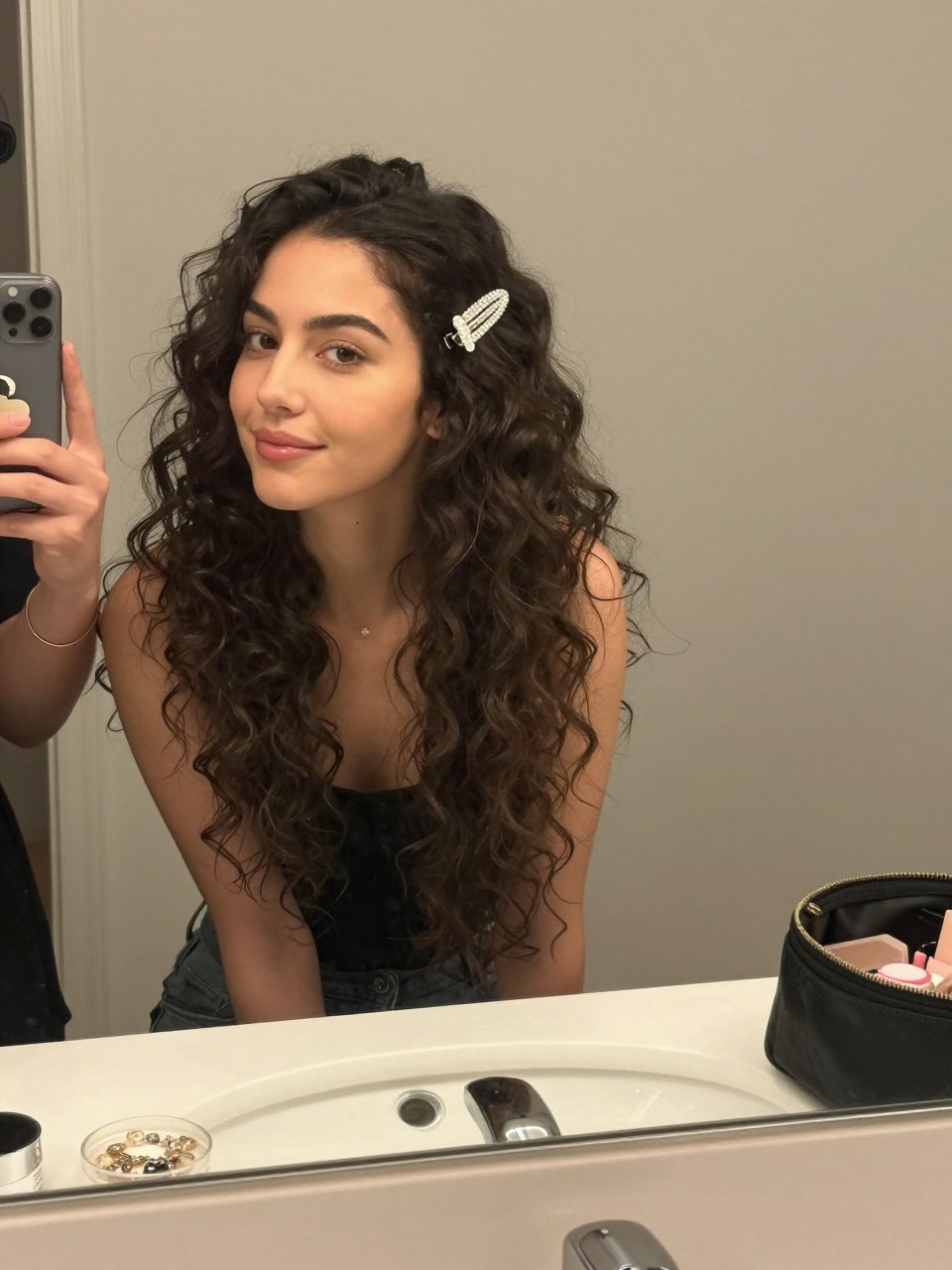 Photo of a woman wearing half up half down curls secured with a crystal clip, candid mirror selfie, setting in a bathroom vanity mirror, lighting soft indoor lighting, containing a small lived-in detail such as a jewelry dish on the counter and a makeup bag nearby, latest iPhone photo quality.