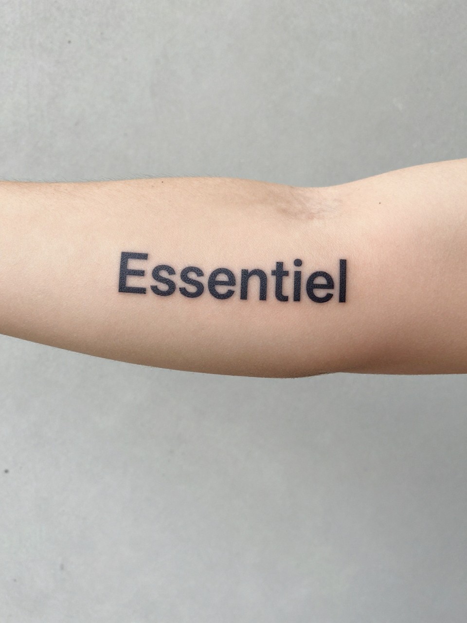  "Close-up of 'Essentiel' in clean, minimalist sans-serif font on the inner bicep. The arm is against a plain, concrete wall. Focus on simplicity and clarity."