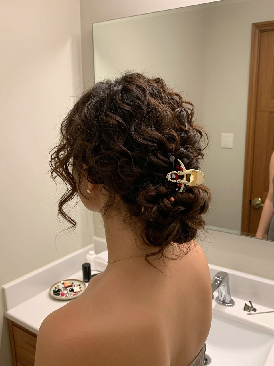 Photo of a woman wearing a curly low bun with a decorative side clip, back-of-head view to show the bun and clip, setting in a bathroom vanity mirror, lighting soft indoor lighting, containing a small lived-in detail such as a jewelry dish on the counter and bobby pins nearby, latest iPhone photo quality.