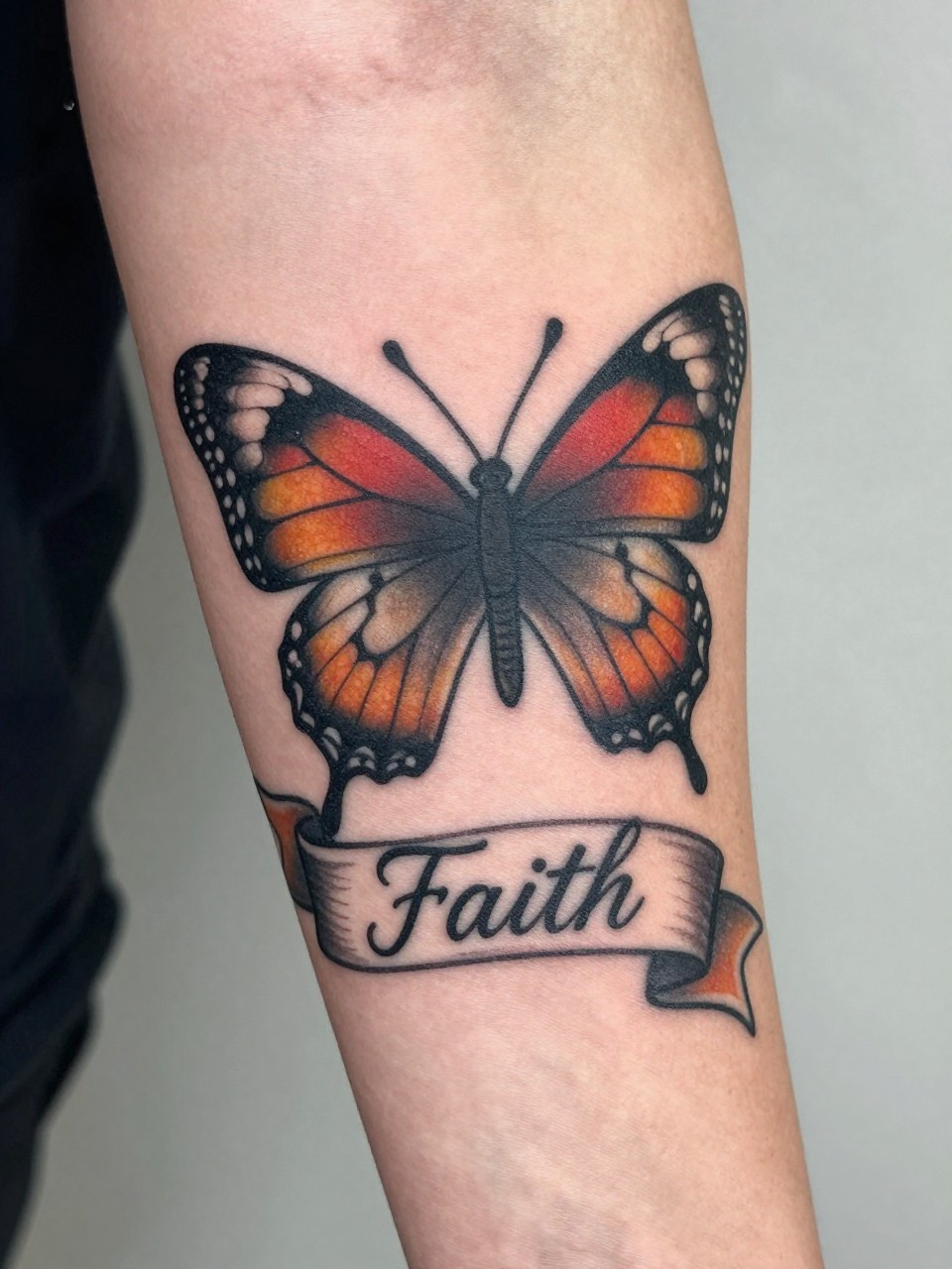  "Detailed view of a traditional butterfly tattoo on the forearm, with a graceful banner beneath it reading "Faith" in cursive. Bold outlines, rich color, and classic elegance."