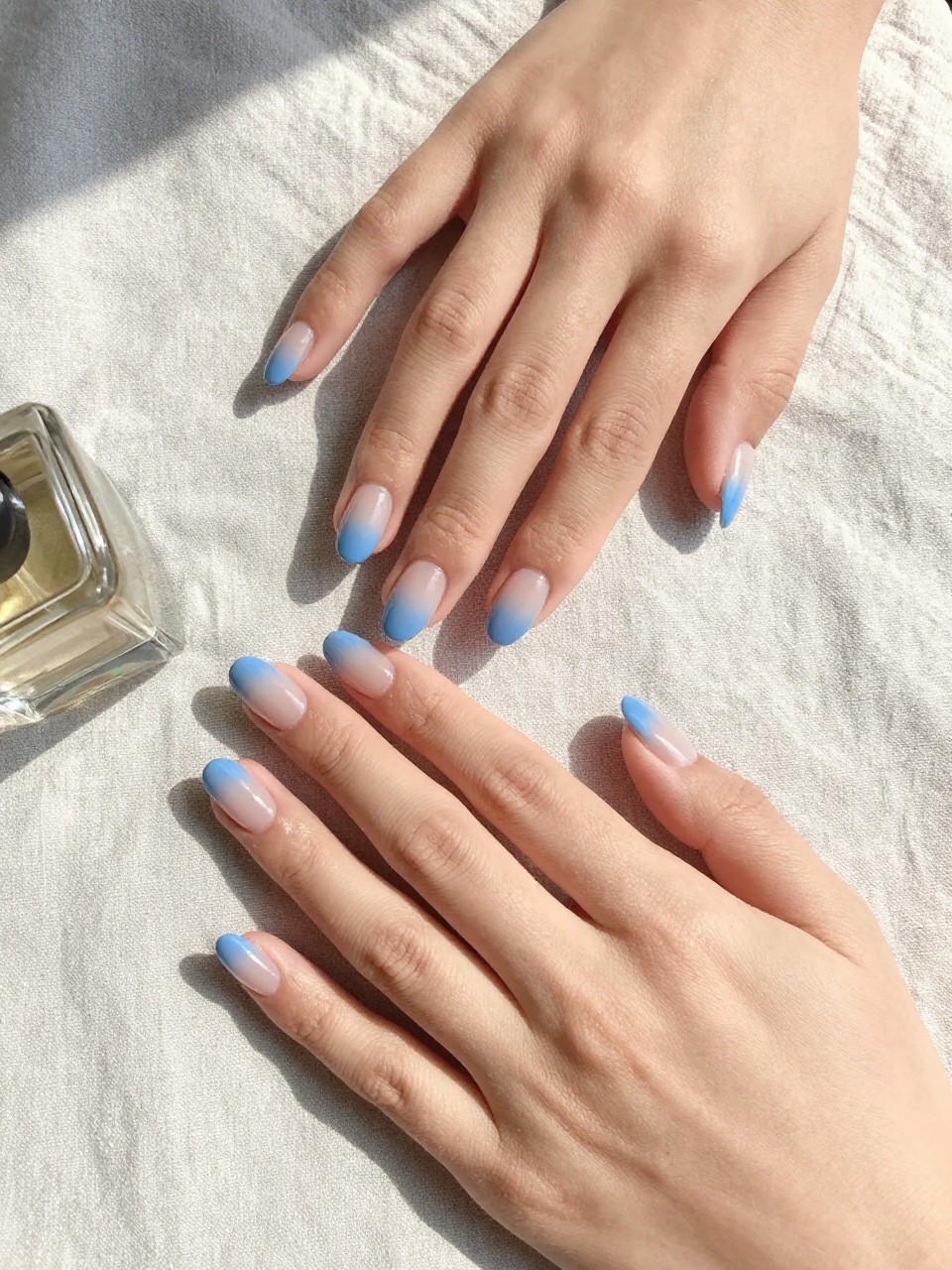 90-degree overhead top-down view of two feminine hands with oval medium nails.
The manicure features a sheer base fading into sky blue tips in a soft ombré blend.
Include glossy gel finish.

Hands are posed one hand resting lightly over the other.

Scene set on a soft linen cloth with a small perfume bottle nearby.

Bright natural daylight with soft shadows, hyper-realistic lifestyle photography, sharp focus on nails, natural skin texture, realistic hands, no extra fingers.